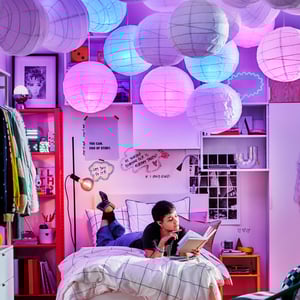 Teenager si čte knihu na posteli pod 18 stínidly závěsné lampy REGOLIT/HEMMA; některá jsou tmavá, jiná svítí růžově nebo modře.