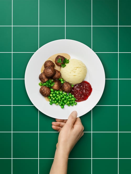 Eine Hand schiebt einen Teller mit HUVUDROLL Proteinbällchen mit Erbsen, Kartoffelpüree und Soße über eine grün karierte Fläche.