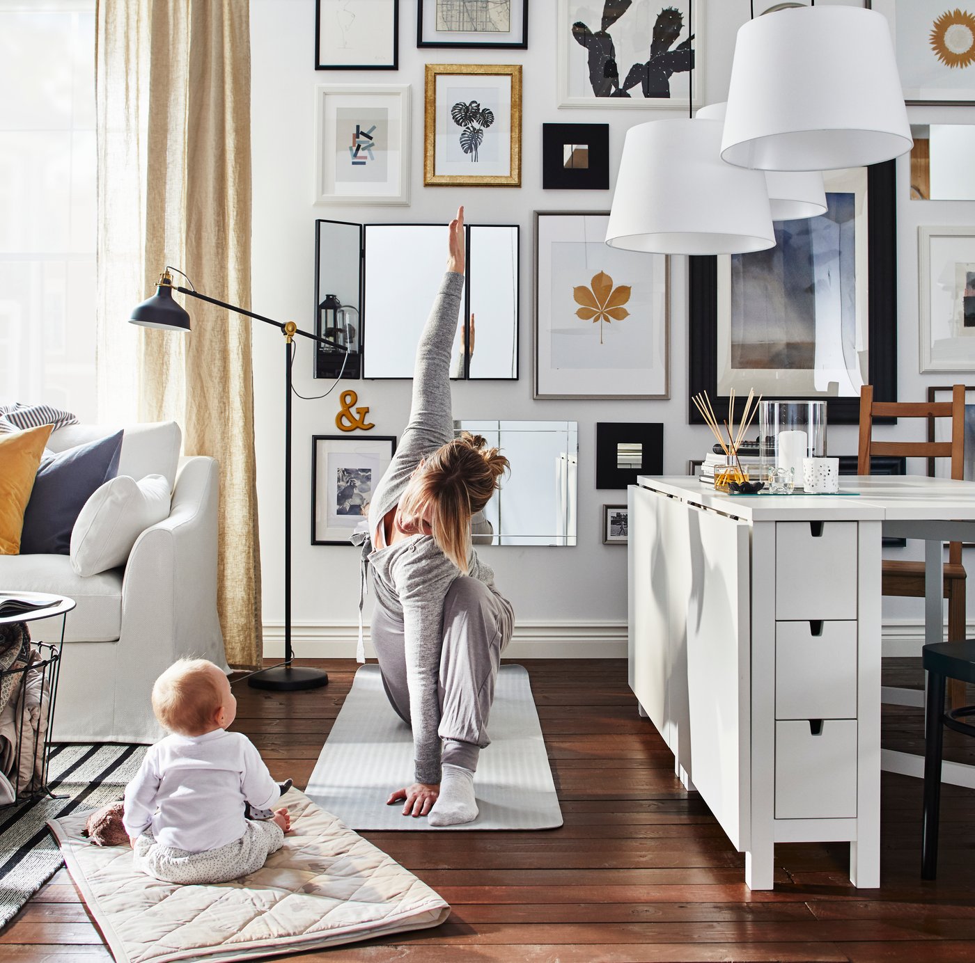 Obývací pokoj s bílou pohovkou, zlatými závěsy a galerií obrazů na stěně. Osoba cvičí jógu na podložce, zatímco malé dítě sedí na měkké podložce. V popředí je bílý úložný stůl IKEA BESTÅ, stojací lampa IKEA RANARP a dekorace včetně rámovaných obrázků a závěsných lamp IKEA.
