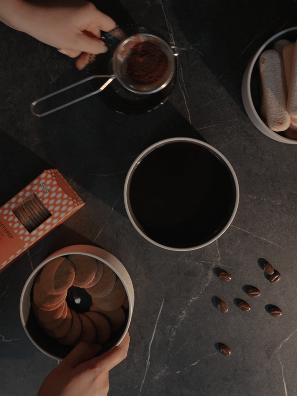 Hände bereiten Kaffee mit einem Sieb über einer schwarzen Tasse auf einer dunklen Oberfläche zu, umgeben von einer runden Dose mit Keksen, Löffelbiskuits und verstreuten Kaffeebohnen. Die Atmosphäre ist gemütlich und einladend.