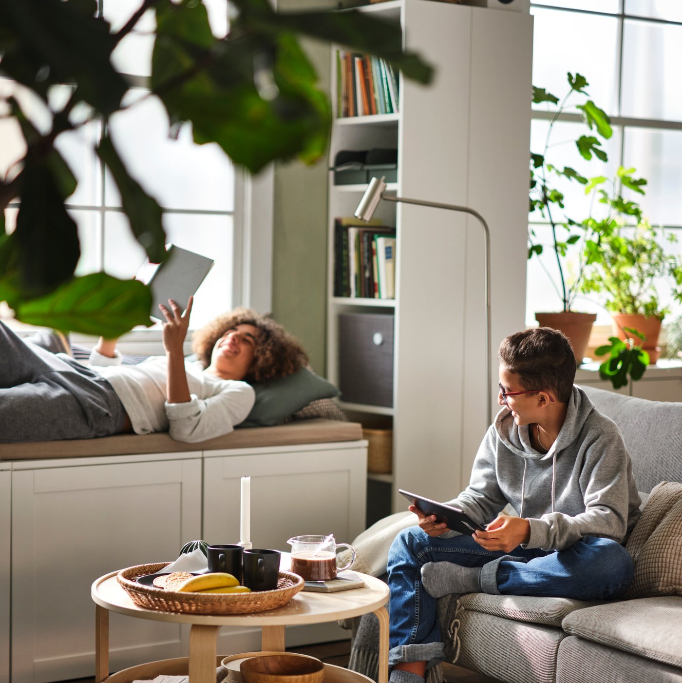 Two teens relaxing in a living area, with one sitting behind a birch veneer BORGEBY coffee table on a SÖDERHAMN 3-seat sofa.