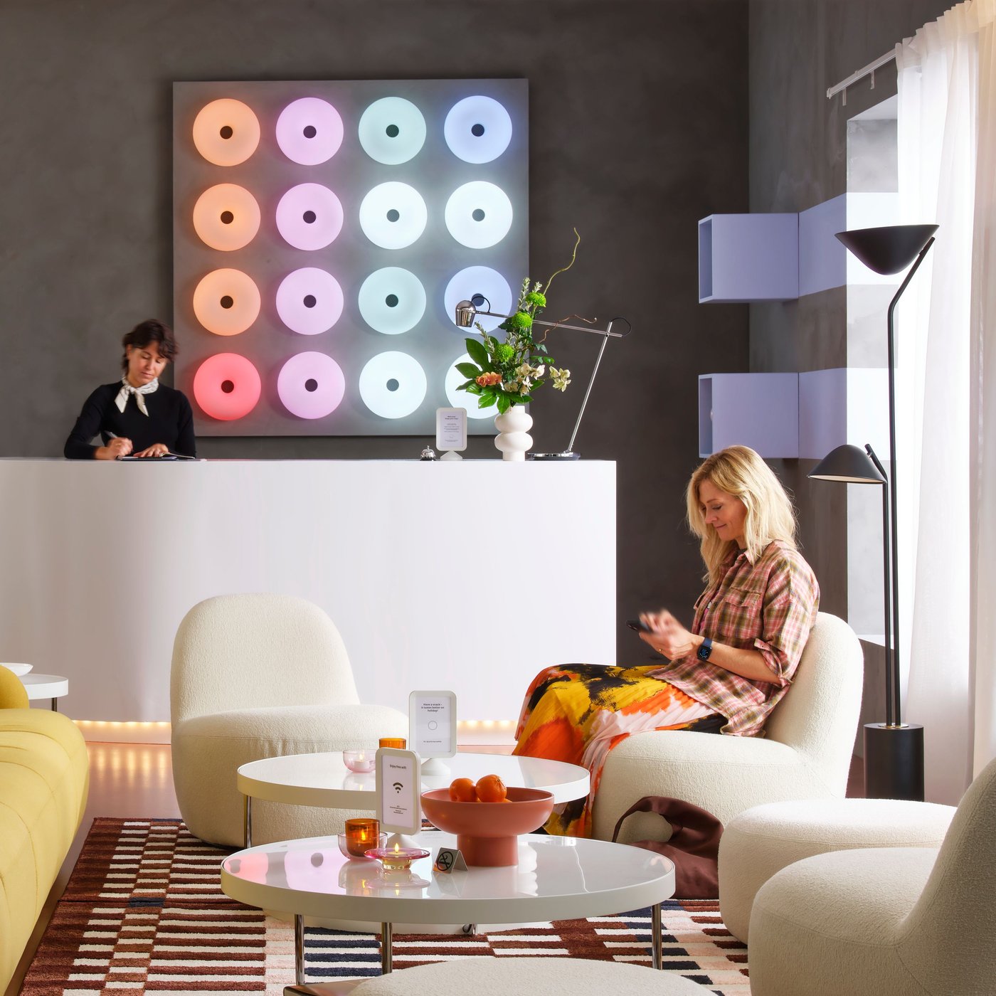 En kvadratisk panel har 16 munkformade VARMBLIXT bord-/vägglampor i olika färger bakom en receptionsdisk i en lounge.