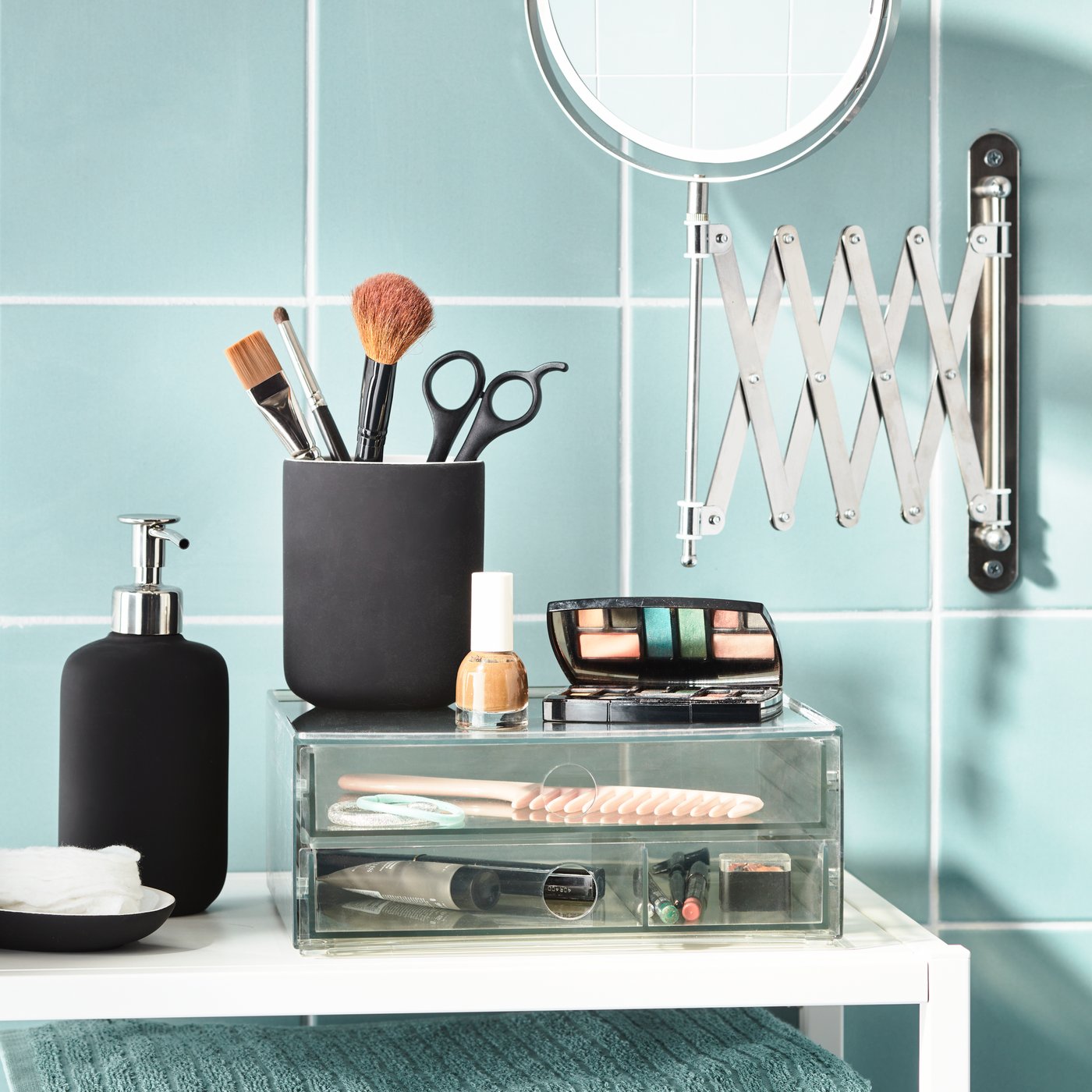 Une élégante composition de salle de bains IKEA présente un distributeur de savon noir, un pot à pinceaux, des cosmétiques, un miroir grossissant et des tiroirs de rangement transparents sur un carrelage turquoise brillant.