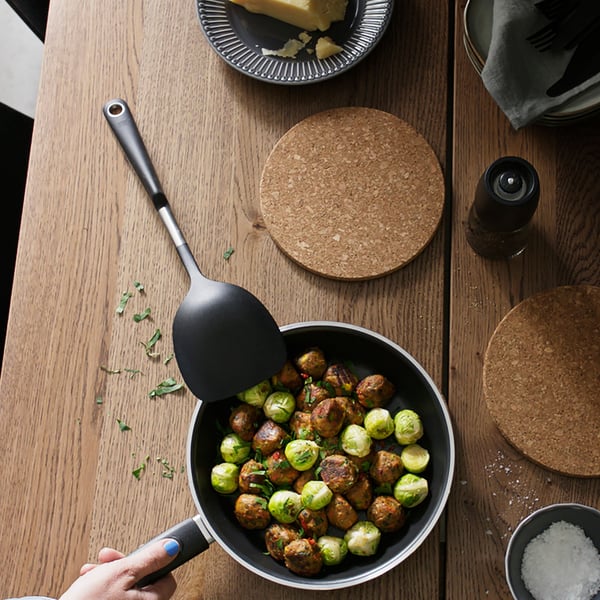 A table with a KAVALKAD frying pan in black containing brussels sprouts and HUVUDROLL plant balls, a HEAT pot stand in cork.