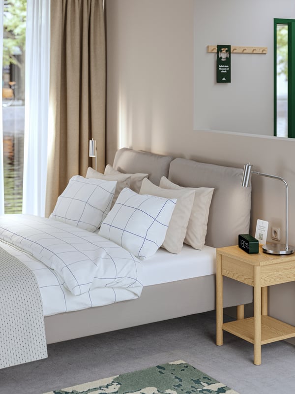 Une chambre d'hôtel avec un lit capitonné SAGESUND réalisé avec une literie BREDVECKLARE à côté d'une table de chevet en placage chêne.