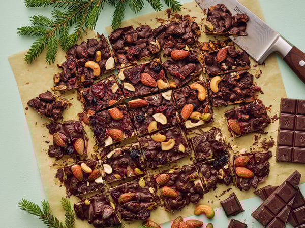 Vista dall'alto di una tavola con scorze di cioccolato con noci e frutti di bosco tagliate in porzioni. Accanto, del cioccolato, un coltello e rami di pino.