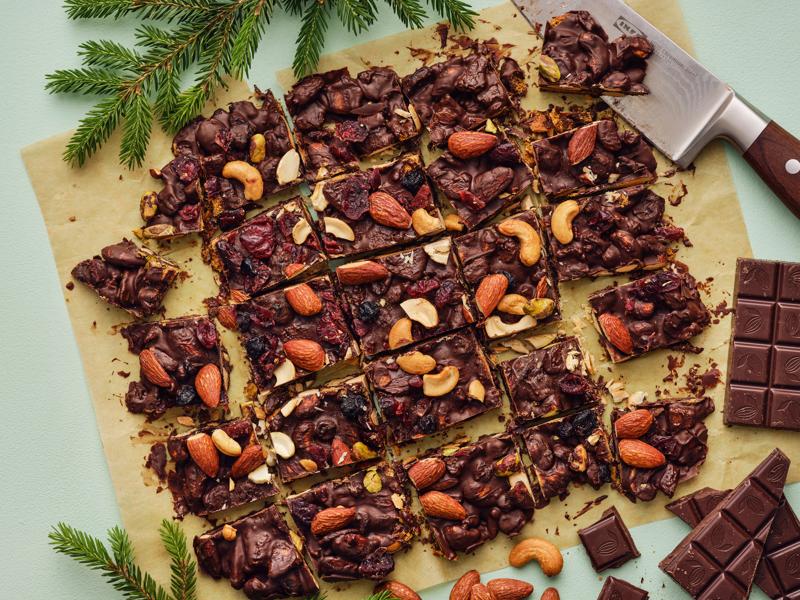 Vista dall'alto di una tavola con scorze di cioccolato con noci e frutti di bosco tagliate in porzioni. Accanto, del cioccolato, un coltello e rami di pino.