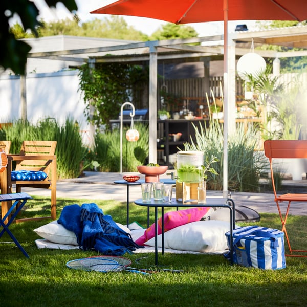 A garden setting with greenery and a sun parasol features a black-blue GRYTSHOLM nest of tables, perfect for outdoor use.