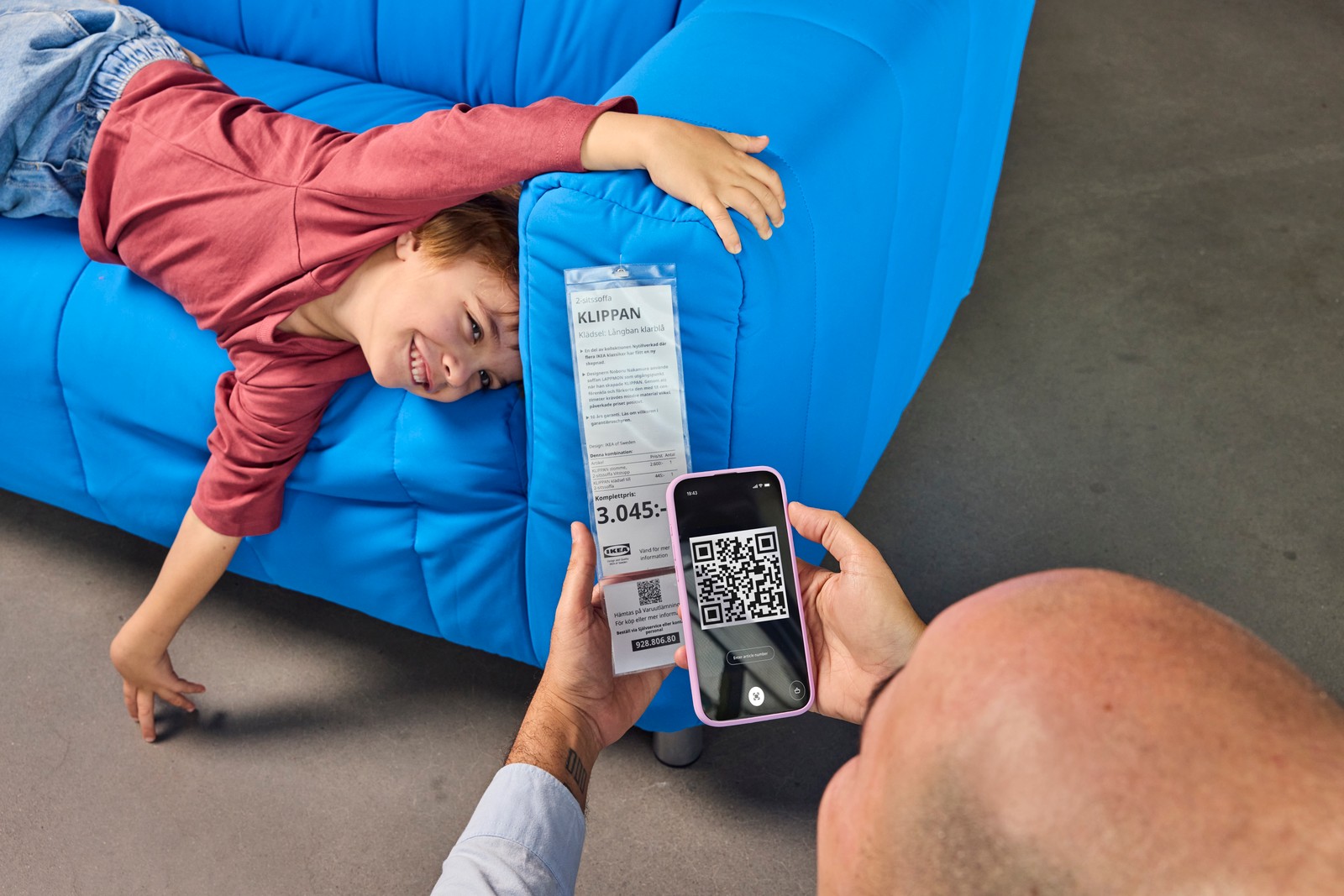 Mężczyzna skanujący kod QR niesbieskiej kanapy, na kórej leży uśmiechnięte dziecko