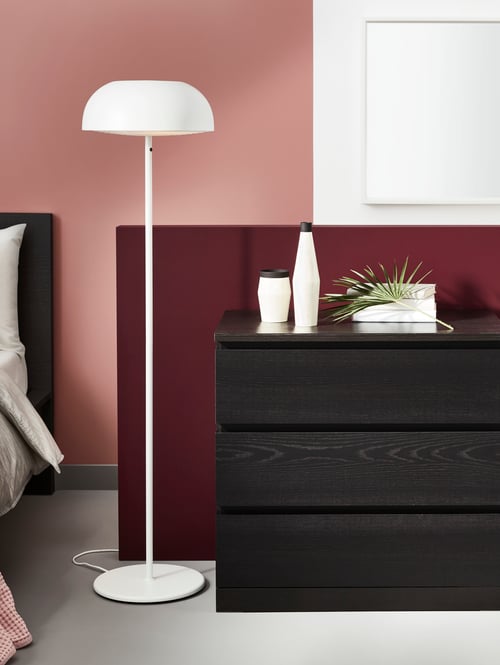 Dormitor cu un dulap cu trei sertare pe care este o lampă și un set de lumânări cu LED, cu o lampă de podea în lateral.