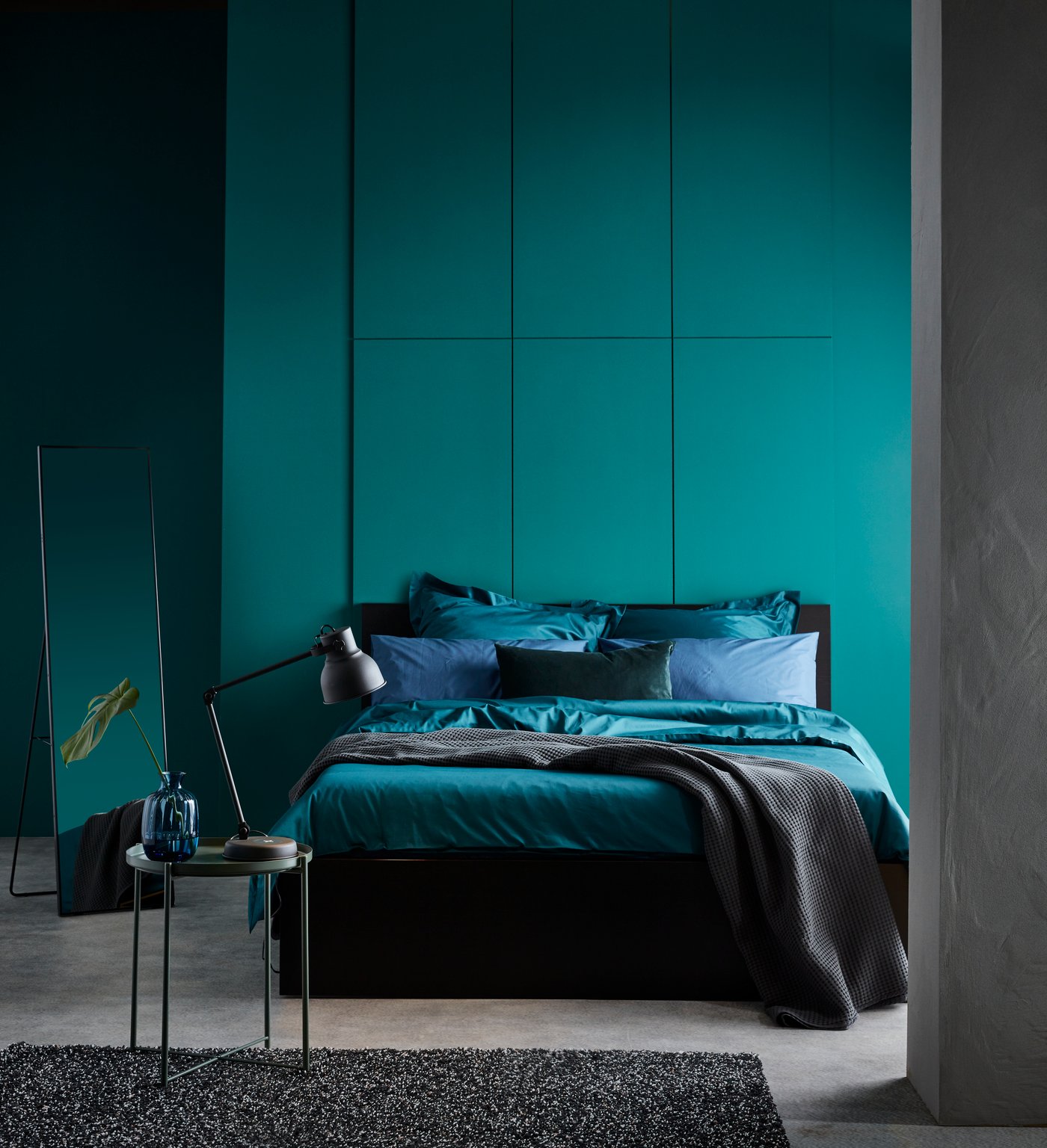 Postel MALM s ložním prádlem, včetně tmavě zeleného povlečení LUKTJASMIN, stojí v ložnici vedle zelené stěny.