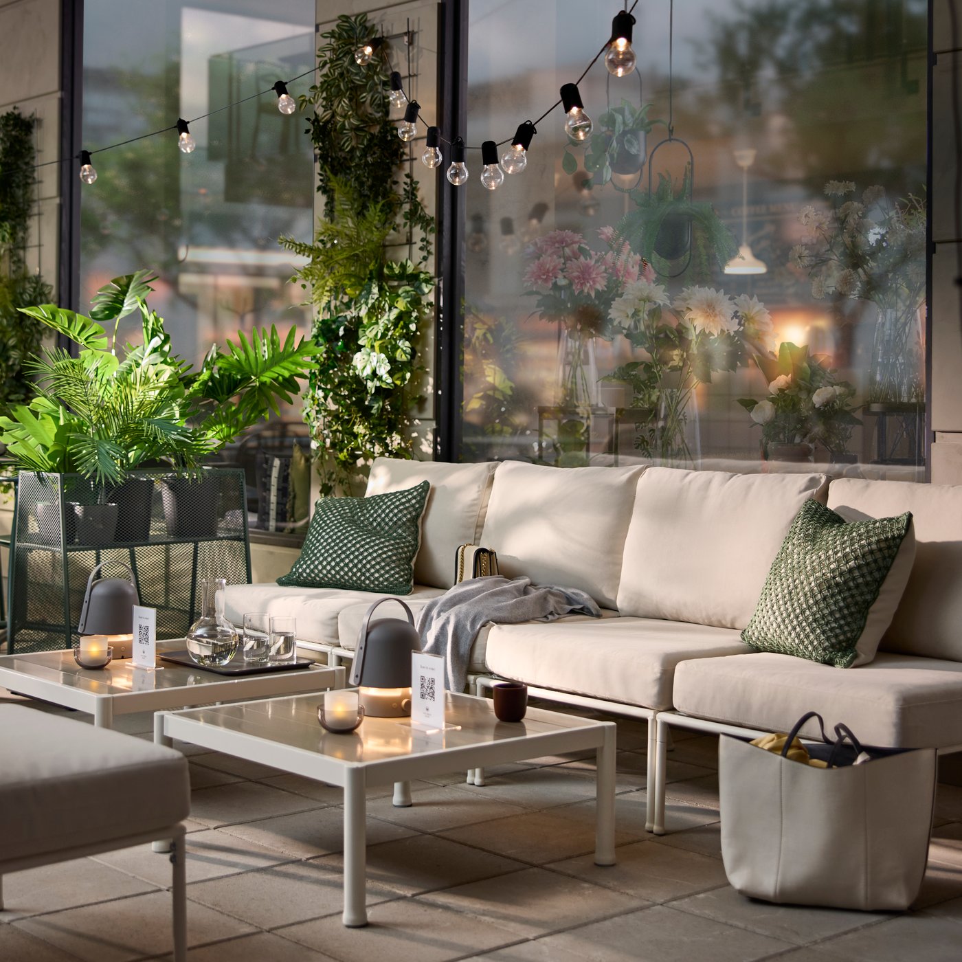 An outdoor café area with a white/beige SEGERÖN seat section for a modular sofa, surrounded by green plants and cosy lights.