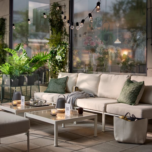 An outdoor café area with a white/beige SEGERÖN seat section for a modular sofa, surrounded by green plants and cosy lights.