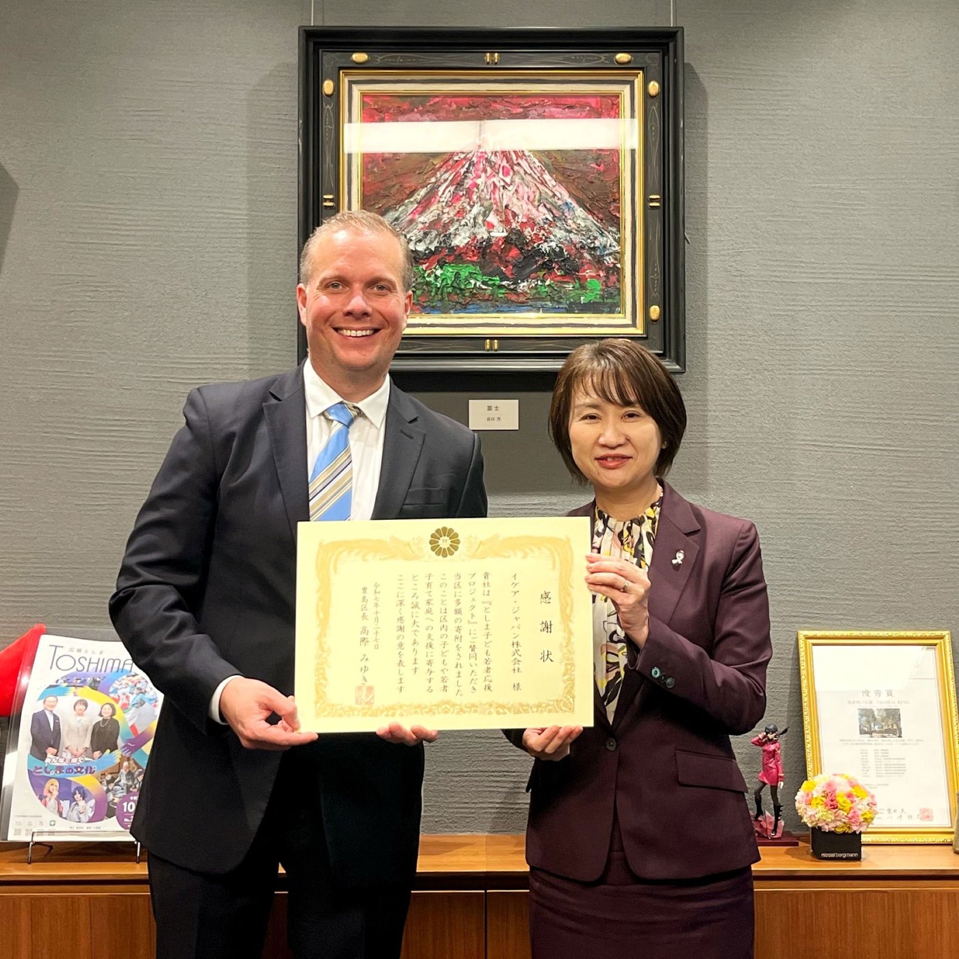 感謝状を手に並ぶ二人。富士山の絵画や表彰状が飾られたオフィスで、受賞の瞬間を記念撮影。