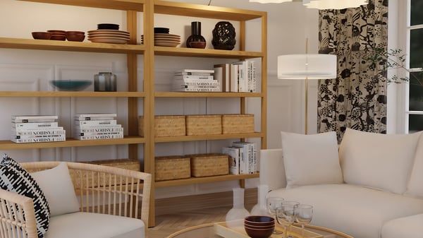 A close‑up of a living room with a light sofa on the right and a rattan armchair with a cushion on the left. In the center stands a round glass coffee table holding small bowls and glasses. Behind the furniture is a tall open shelving unit in light wood filled with books, bowls and baskets. 