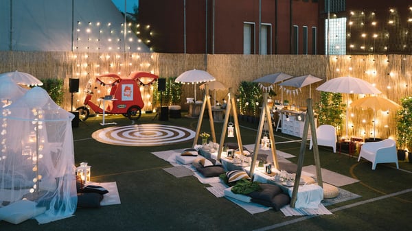 Uno spazio esterno sul far della sera illuminato da lucine decorative e lanterne. Al centro, una tavola bassa allestita e apparecchiata. Attorno, ombrelloni, pouf e cuscini.