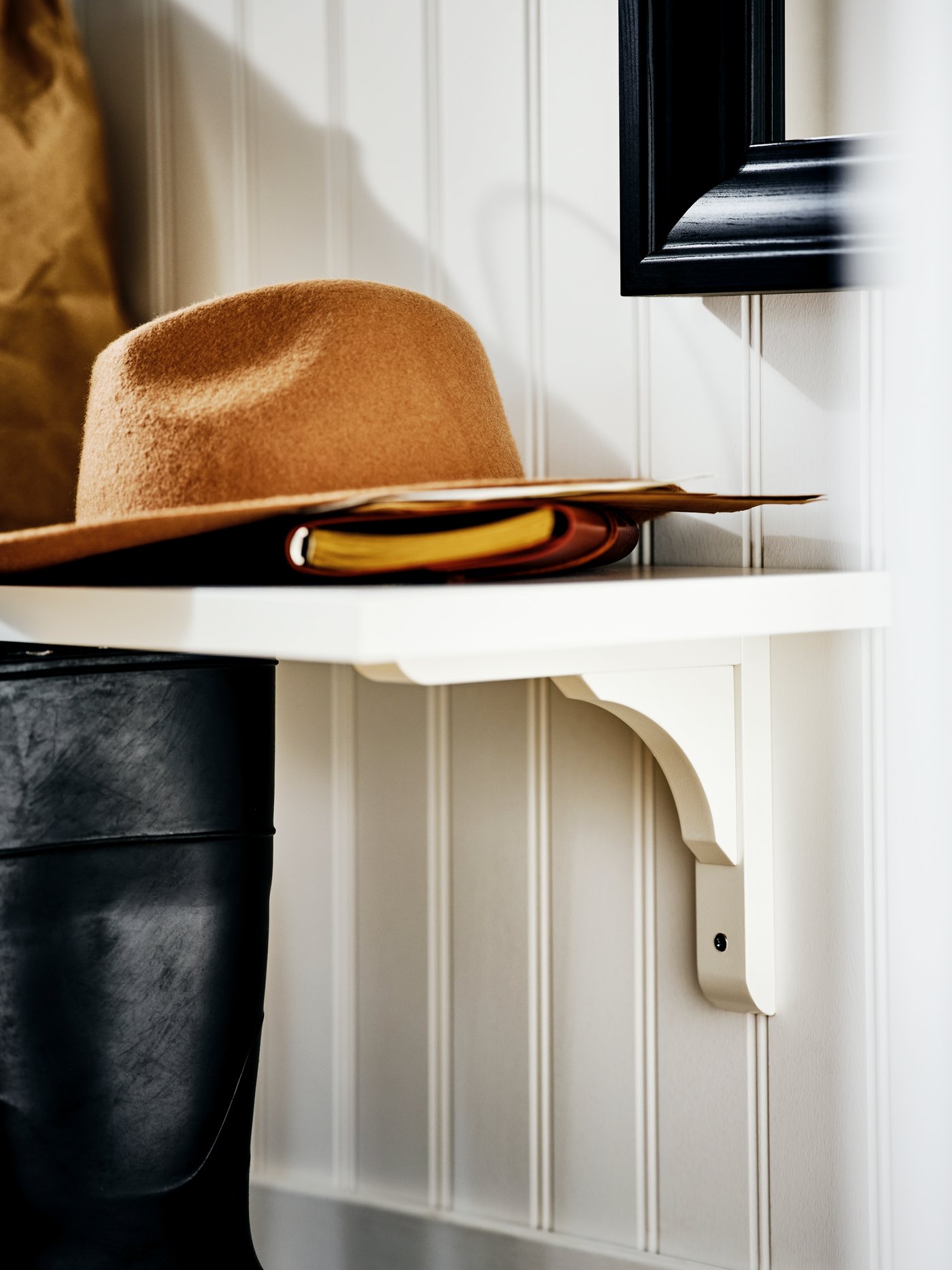 طرف رف جداري BERGSHULT/RAMSHULT أبيض على جدار بألواح خشبية مع قبعة بني دفتر ملاحظات عليه.