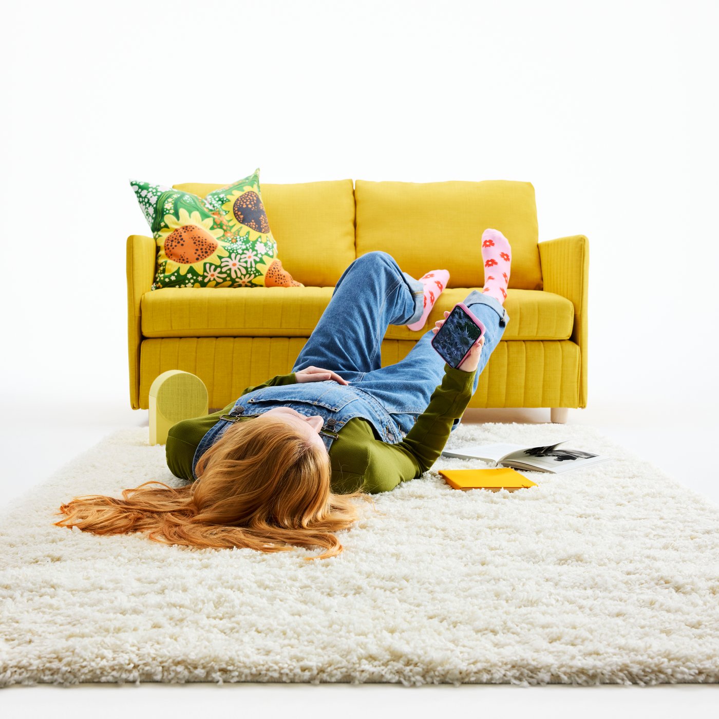 A young woman is relaxing on an off-white ÄRENDE rug, with her feet up on a yellow sofa.