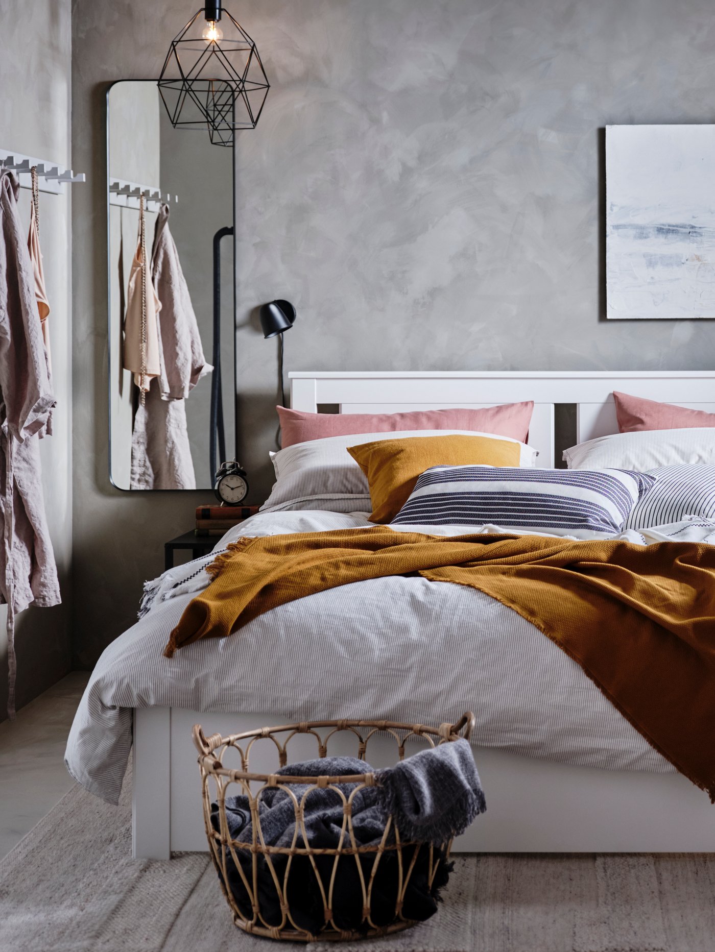Dormitorio con una funda nórdica gris y fundas de almohada sobre una estructura de cama blanca, cojines, un espejo rectangular y una cesta.
