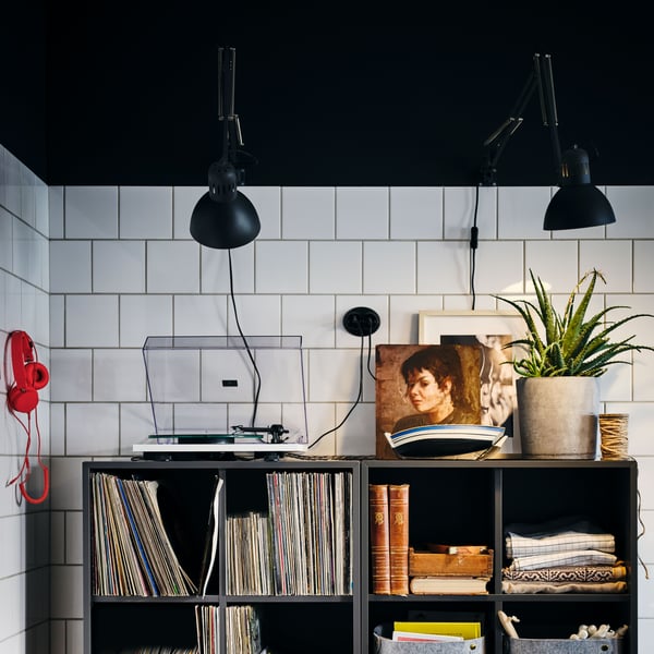 A tiled wall with two black TERTIAL wall spots shining on a record player, a plant, and a photo frame.