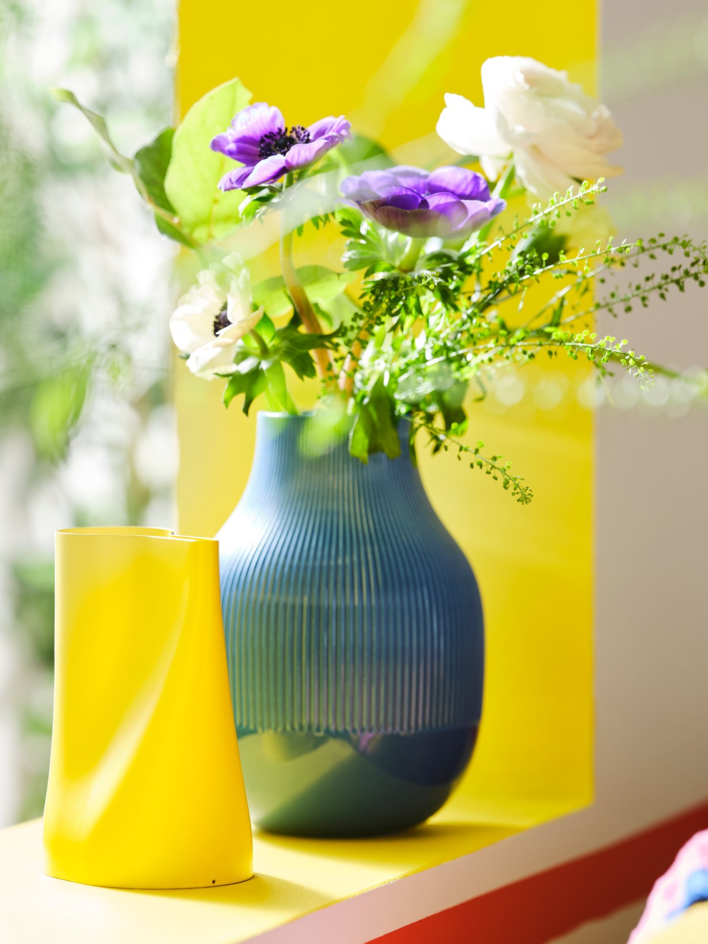 Et vindueshjørne med en blå GRADVIS vase fyldt med blomster og en gul CHILIFRUKT vase.