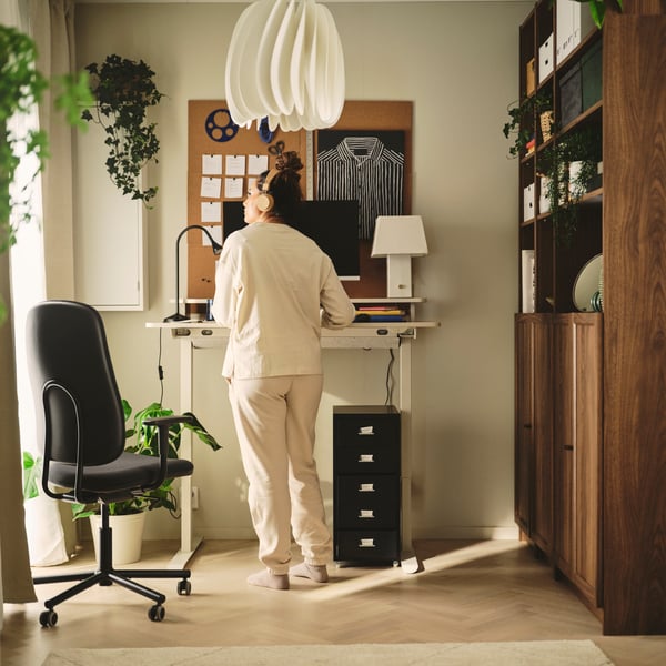 TRAVSPORT Sitz-/Stehschreibtisch in Eichenoptik mit schwarzem Rollstuhl und Zimmerpflanzen im Homeoffice.