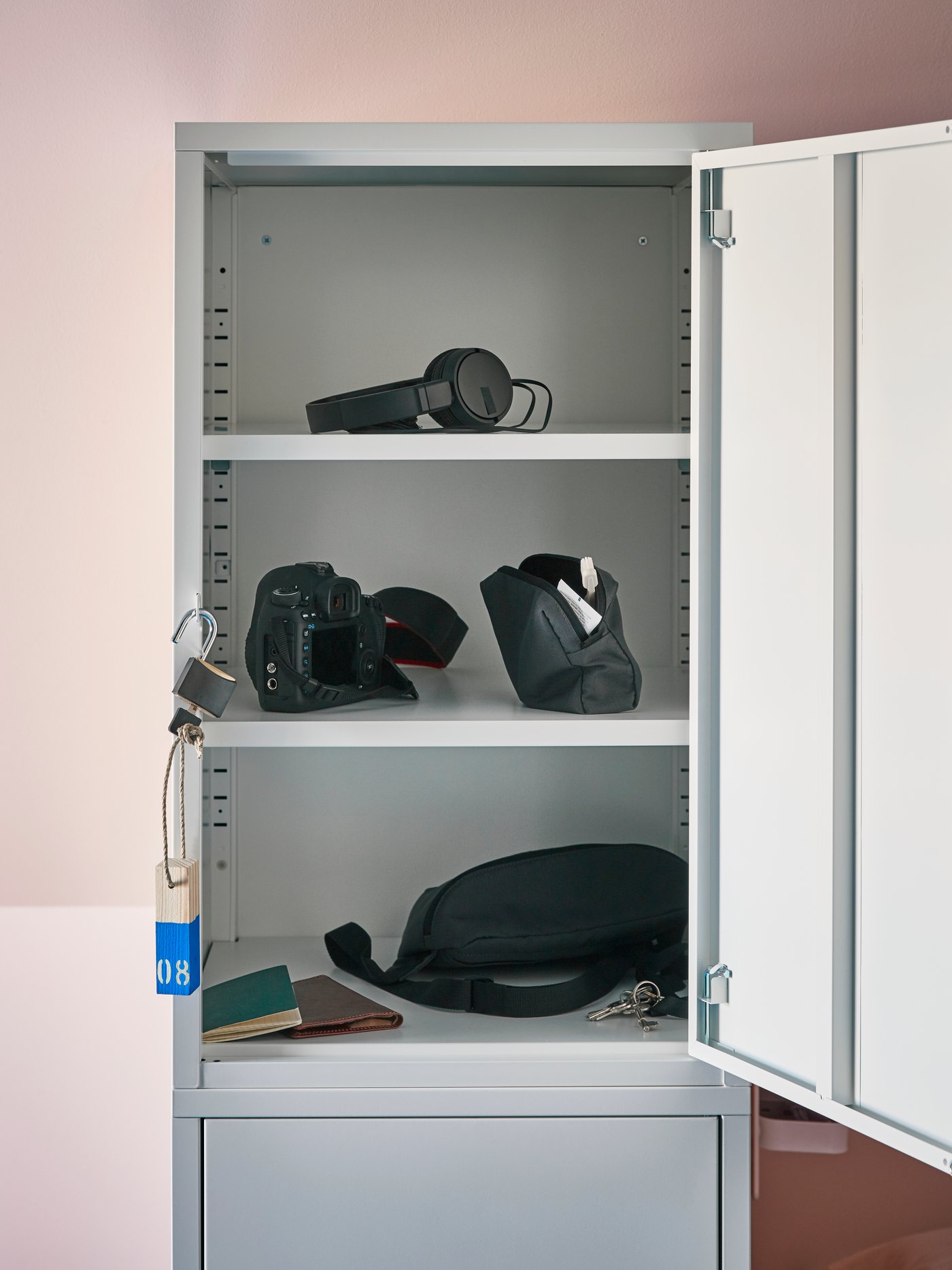 خزانة تخزين ‪HÄLLAN‬ مع باب مفتوح يُظهر سماعات رأس وكاميرا ومتعلقات سفر أخرى على رفوف مختلفة.