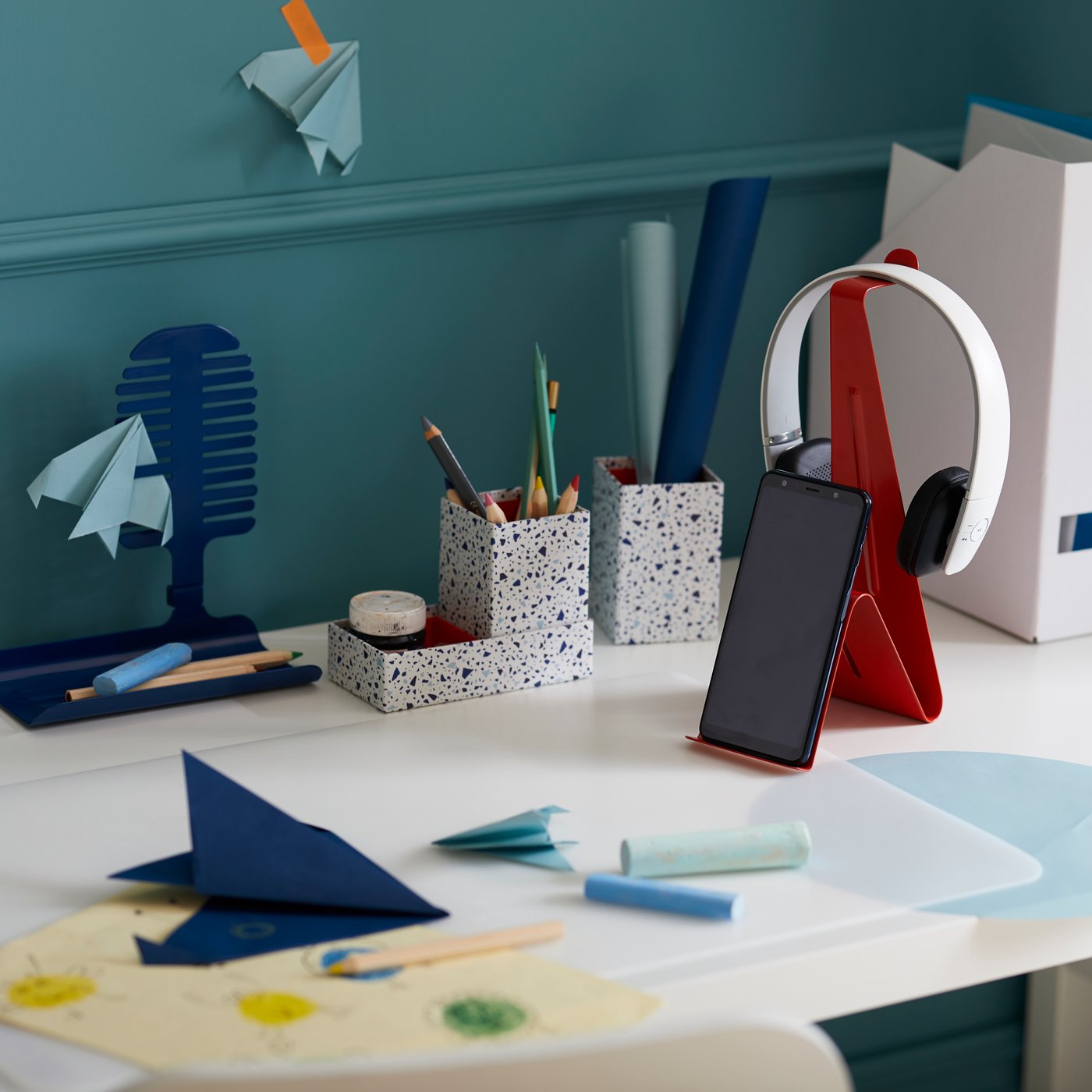 A folded piece of navy blue paper sitting on a table mat atop a white table with a red phone stand supporting a phone and a pair of headphones with stationery holders and a magazine file behind it.