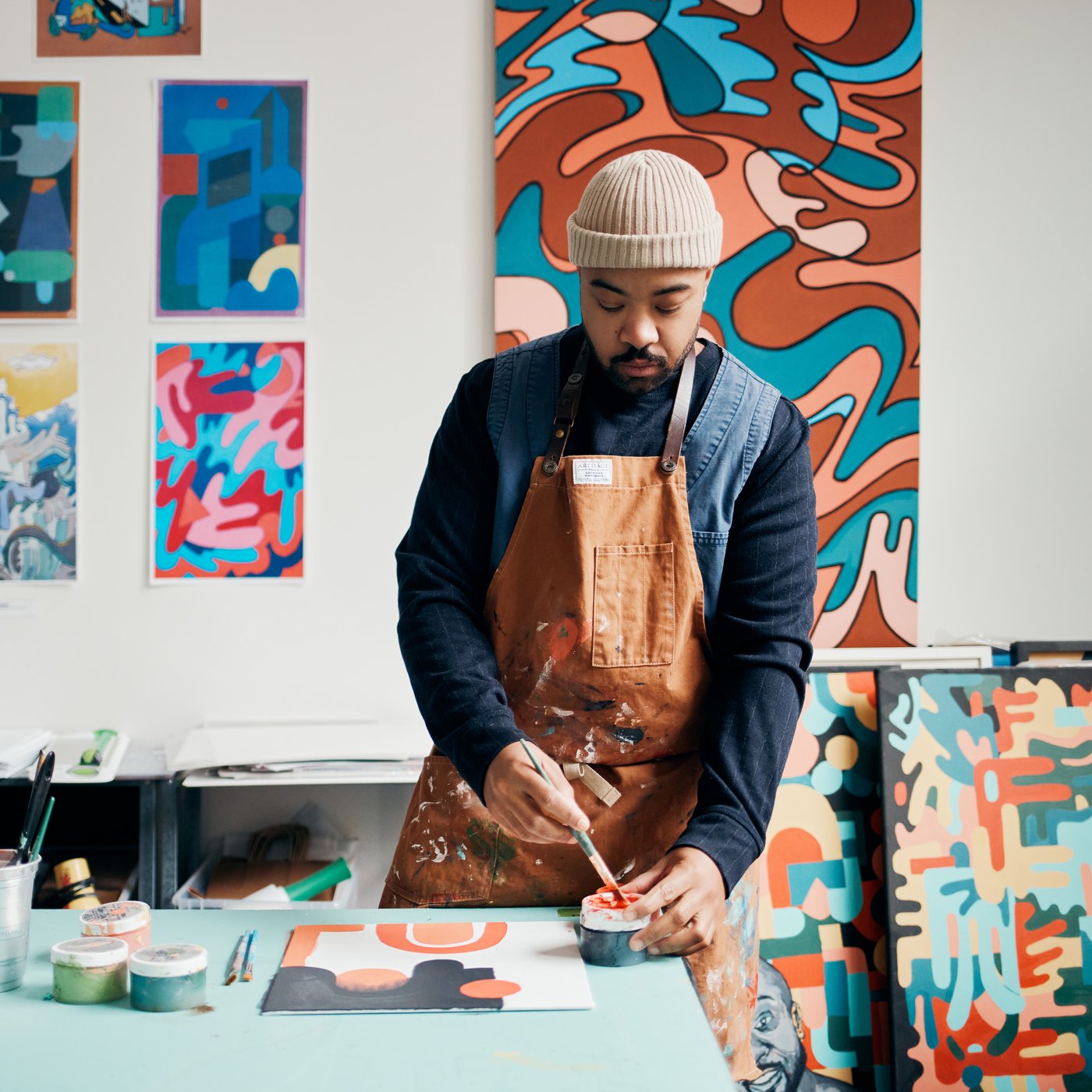 A man in his studio is painting. Around him on the walls are more paintings. The desk is full of artistic supplies.