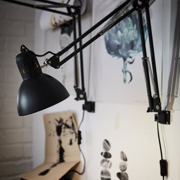 A close-up of a TERTIAL work lamp in dark grey attached to a wall with illustrations in the background.