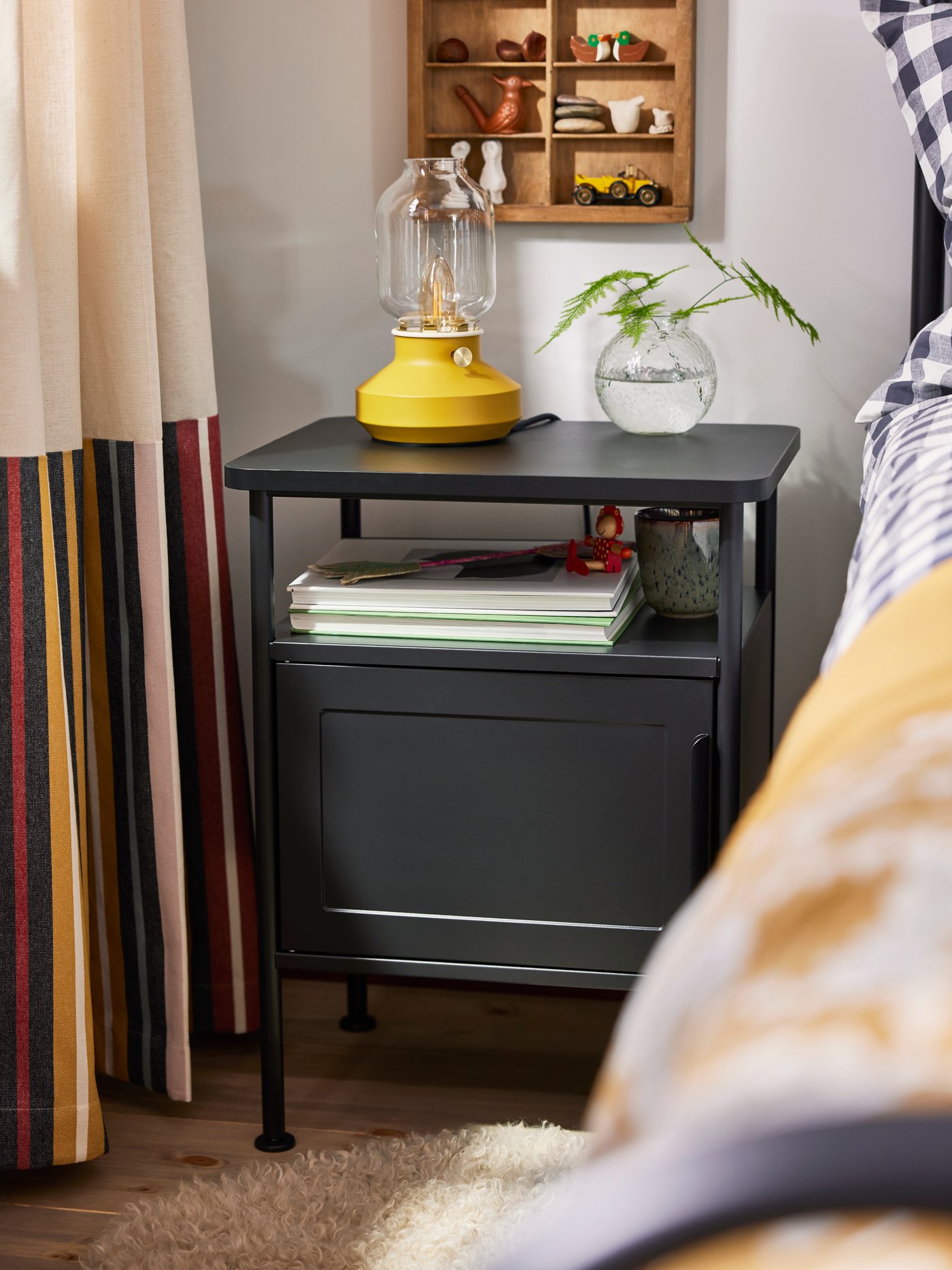 Close-up of GRÅFJÄLLET bedside table in anthracite with yellow TÄRNABY table lamp and small glass vase on top.