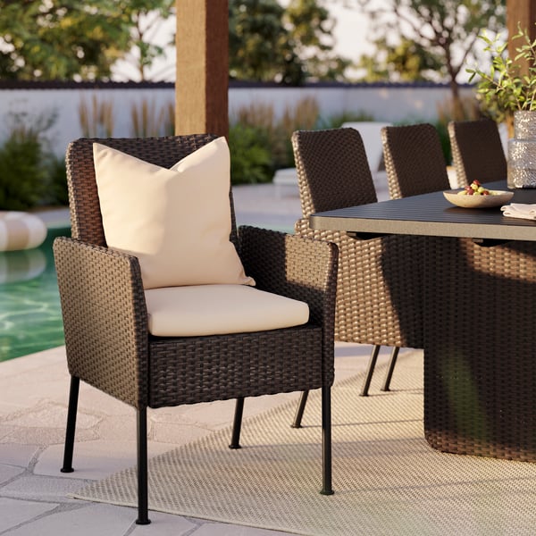Close-up of a VITTSKÄR dining chair in dark grey plastic rattan with two beige pillows. A table and chairs, outdoors.