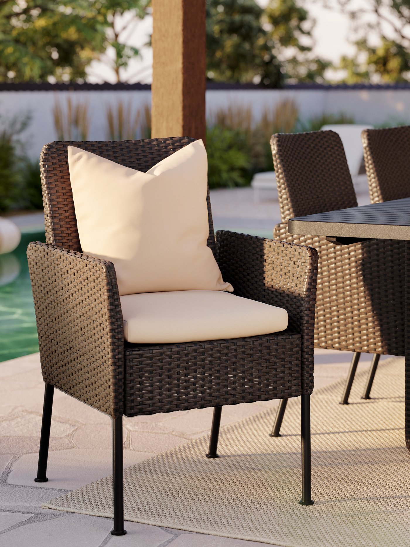 Close-up of a VITTSKÄR dining chair in dark grey plastic rattan with two beige pillows. A table and chairs, outdoors.