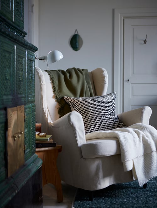Une housse de coussin KUSTFLY noir et beige est posée sur un fauteuil STRANDMON blanc avec des jetés ODDRUN moelleux.