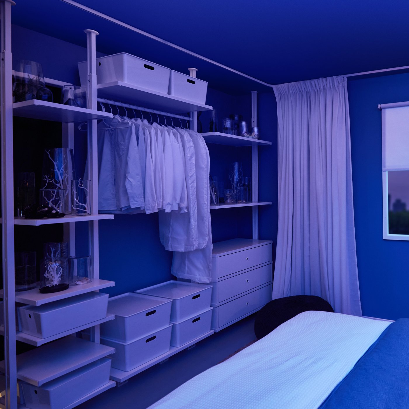 A view of the whole shelving unit in a room with blue lighting. There are white shirts on hangers and a few drawers in the bottom of the unit.
