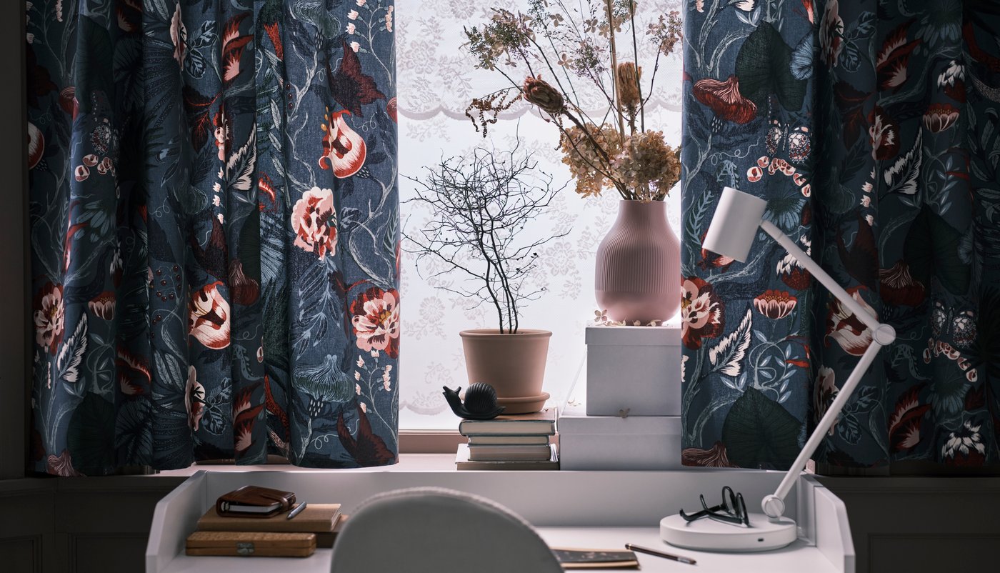 Tende realizzate con tessuto FILODENDRON blu scuro/motivo floreale appese a una finestra sopra una scrivania HAUGA bianca con una lampada da lavoro NYMÅNE bianca.