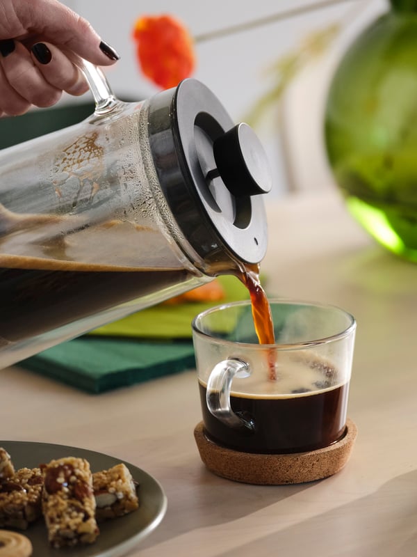 Coffee being poured from a coffee pot into a mug on a cork coaster. In the background is a green vase and orange flower.