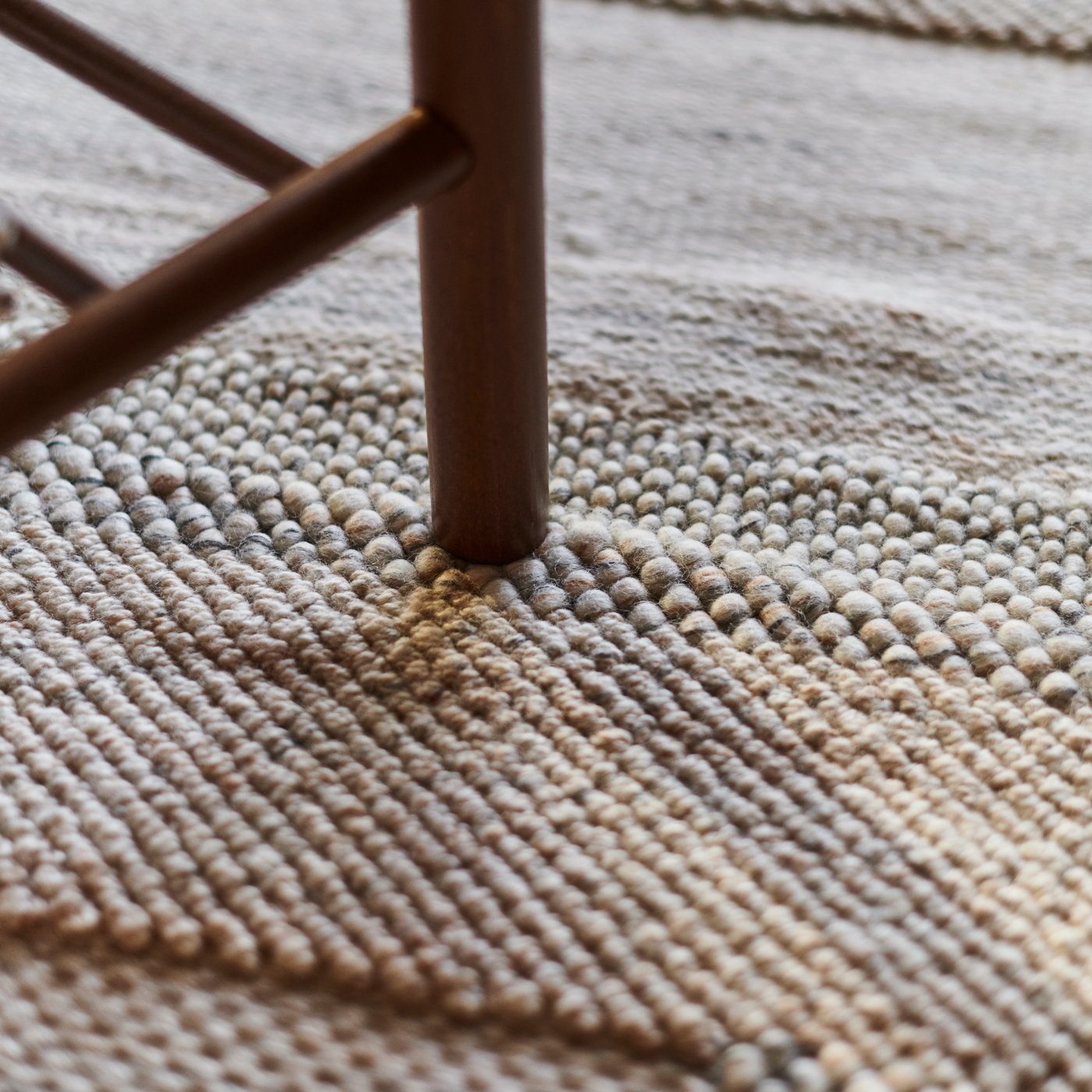The different textures of a light-coloured BRÖNDEN low pile rug on the floor with a dark brown chair leg on it.