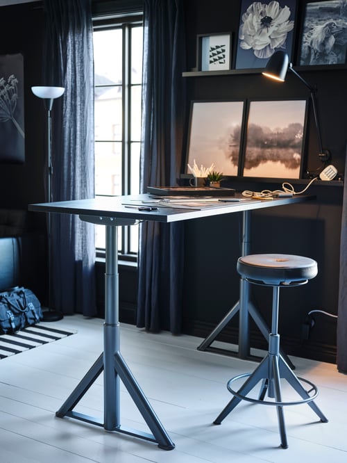 A desk against a wall which has black-framed photos on it, with a black TROLLBERGET active sit/stand support in front of it.