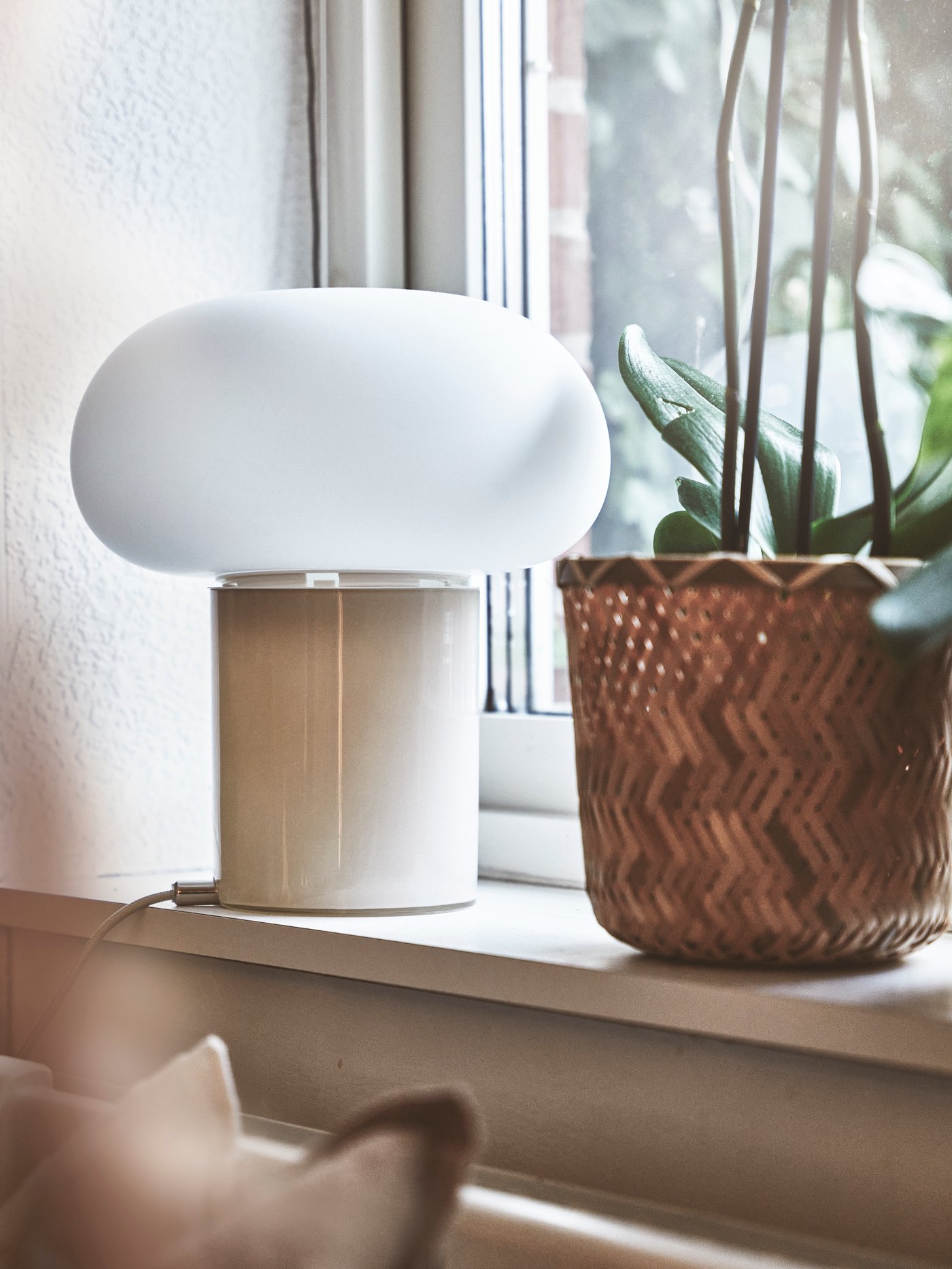 Vit bordslampa med rund, opak skärm står på en fönsterbräda bredvid en krukväxt i en flätad korg. Fönstret i bakgrunden släpper in ljus.