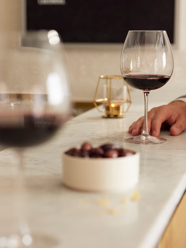 Una mano appoggiata a un bancone. Vicino dei bicchieri  STORSINT con del vino rosso. Sul piano, una ciotola con del cibo e un portacandela PÄRLBAND.