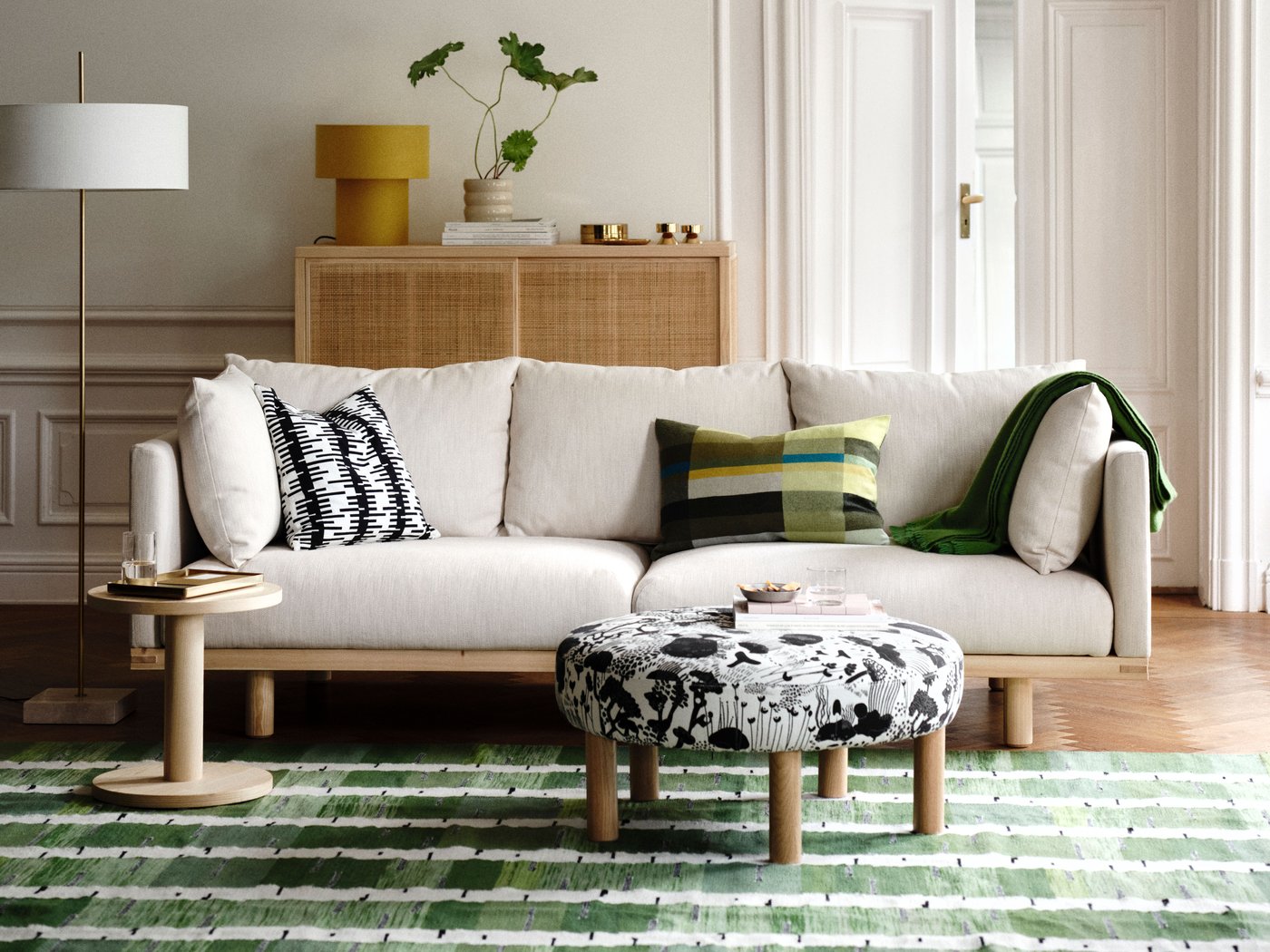 Ett ljust och modernt vardagsrum med en beige soffa med prydnadskuddar, en mönstrad fotpall och en grönrandig matta, allt arrangerat kring trämöbler.