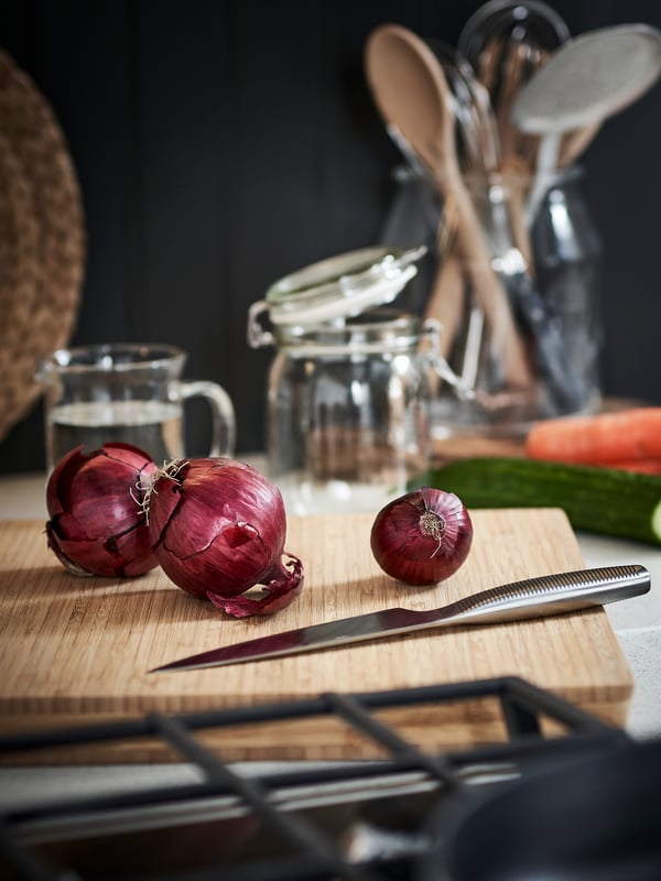 In einer Küche sieht man ein Schneidbrett auf dem rote Zwiebel und ein IKEA 365+ Gemüsemesser liegt. Dahinter sieht man ein leeres KORKEN Glas und eine BEGÄRLIG Vase in der Küchenutensilien verstaut sind.