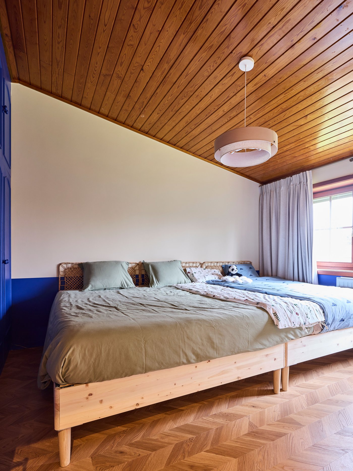 Slaapkamer met houten bed, beige beddengoed, schuin houten plafond en hanglamp, met raam en gordijnen voor warm, rustgevend interieur.