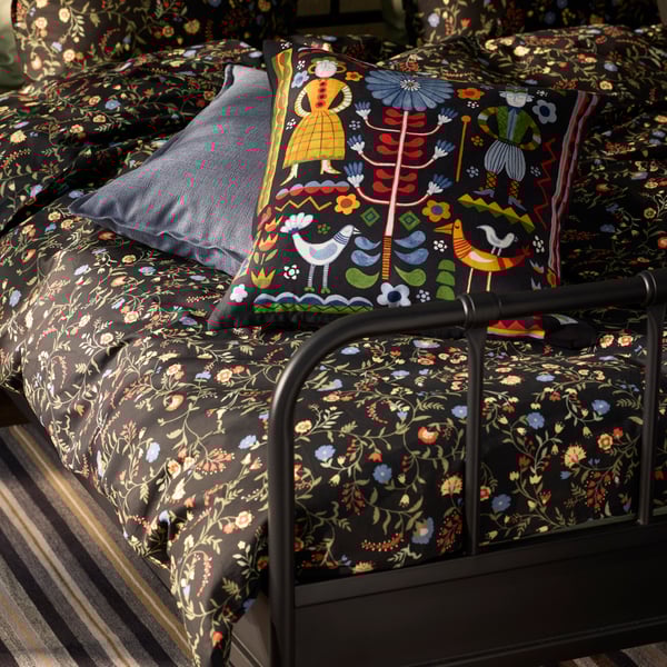 A floral BRUDKRUSBÄR duvet cover and pillowcase complemented by a colourful TRATTKAKTUS cushion cover on a dark bed frame.