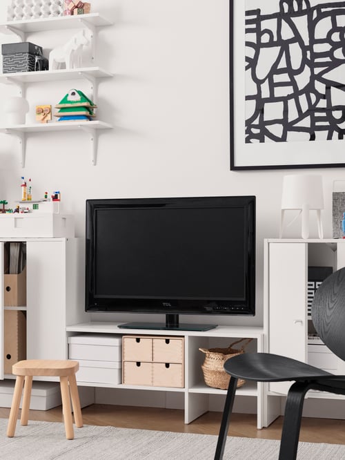 Un salon dans lequel on retrouve un meuble télé blanc, un téléviseur, une armoire avec porte ouverte, un fauteuil relax noir, des tablettes murales et un tabouret pour enfants.