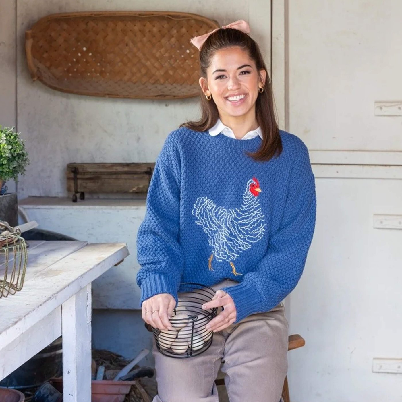 Person in blue sweater with chicken design sits holding a wire basket in a rustic kitchen.
