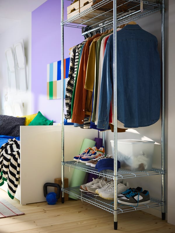 A steel OMAR shelving unit with clothes rail standing at the foot end of a bed, with shoes, clothes and boxes neatly stored.