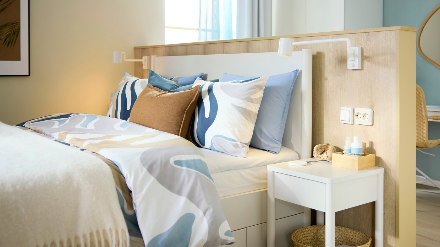 A bed has a leafy-patterned MYGGLASVINGE quilt cover and pillowcases on it, against a low wall in a light-filled hotel room.