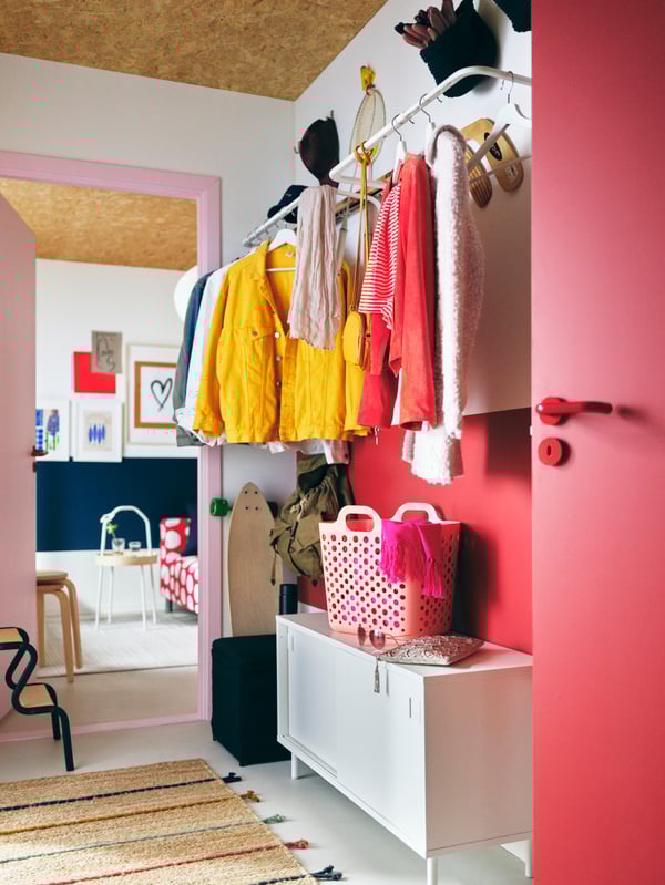 A hallway in reds and whites with garments on white MULIG clothes bars on the wall above a white MACKAPÄR storage bench.
